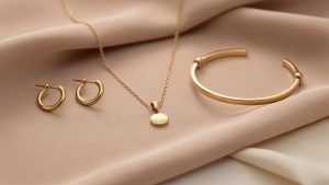 Composition élégante d’un collier fin, de créoles et d’un bracelet doré disposés sur un fond beige poudré, éclairée par une lumière douce.